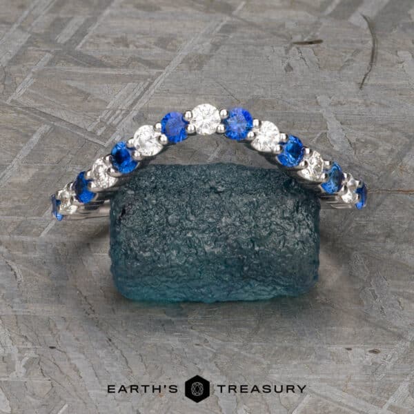 The Champlain band in 18k white gold The "Champlain" Band, a curved silver ring adorned with alternating blue and white gemstones, is displayed upright on a textured gray surface. The round stones are evenly spaced, creating a striking contrast. The brand name "Earth's Treasury" is visible below the ring.