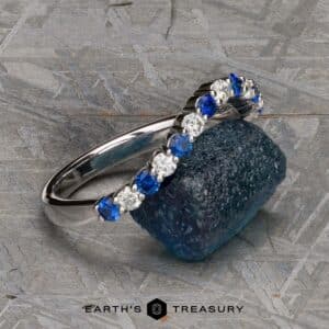 A silver "Champlain" Band adorned with alternating blue and white gemstones rests on a deep blue stone. The image has a textured, gray background and features the logo and text "Earth's Treasury" at the bottom center.