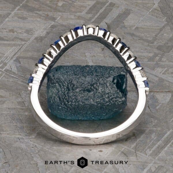 The Champlain band in 18k white gold The "Champlain" Band is an elegant silver ring adorned with a series of blue gemstones embedded along the top, displayed upright on a textured surface with a greenish-blue stone placed behind it. The bottom portion of the ring displays the words "EARTH'S TREASURY.