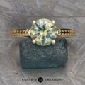 The "Kerria" Solitaire ring is showcased on a blue textured mineral. This exquisite piece features a round-cut, clear gemstone securely held by six prongs on a gold band with an intricate patterned design. The ring stands out against a rough-textured gray background, with the text "EARTH'S TREASURY" displayed at the bottom.