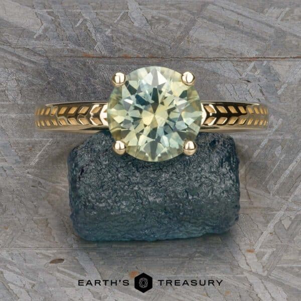 The Kerria Ring in 14k yellow gold with 2.28-carat Montana sapphire The "Kerria" Solitaire ring is showcased on a blue textured mineral. This exquisite piece features a round-cut, clear gemstone securely held by six prongs on a gold band with an intricate patterned design. The ring stands out against a rough-textured gray background, with the text "EARTH'S TREASURY" displayed at the bottom.