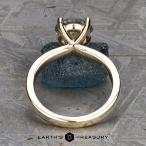 The "Kerria" Solitaire ring, set with a light green gemstone in an elegant V-shaped prong setting, features a sleek, modern design. Displayed upright on a textured gray surface, it is accompanied by the brand name "Earth's Treasury" below.
