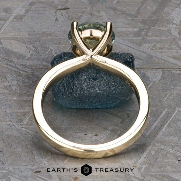 The Kerria Ring in 14k yellow gold with 2.28-carat Montana sapphire The "Kerria" Solitaire ring, set with a light green gemstone in an elegant V-shaped prong setting, features a sleek, modern design. Displayed upright on a textured gray surface, it is accompanied by the brand name "Earth's Treasury" below.