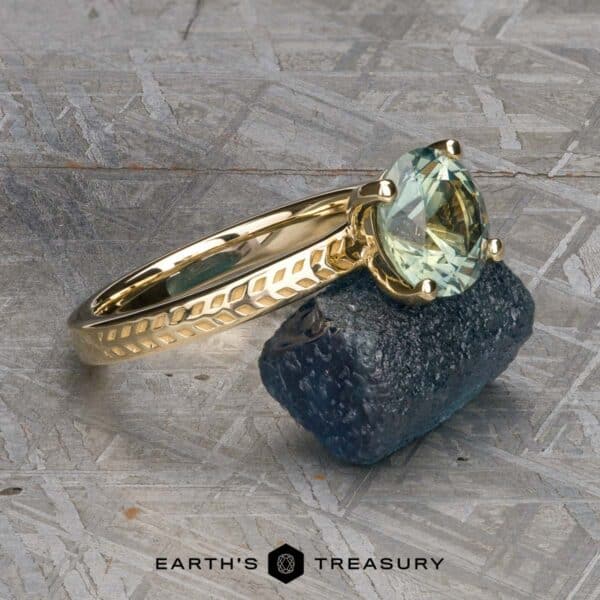 The Kerria Ring in 14k yellow gold with 2.28-carat Montana sapphire The "Kerria" Solitaire, a stunning gold ring adorned with a green oval gemstone that could be aquamarine, lies elegantly on a textured gray surface. The band of the ring showcases intricate engraved patterns and is placed atop a piece of rough blue stone. At the bottom, the text "EARTH'S TREASURY" is prominently displayed.