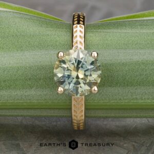 A round-cut light yellow-green gemstone set in a gold ring with a patterned band, displayed on a green leaf background. The name "The 'Kerria' Solitaire" is visible at the bottom of the image.