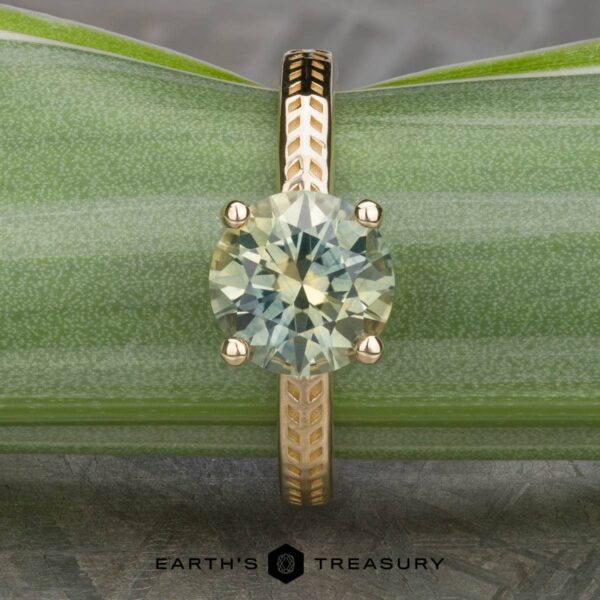 The Kerria Ring in 14k yellow gold with 2.28-carat Montana sapphire A round-cut light yellow-green gemstone set in a gold ring with a patterned band, displayed on a green leaf background. The name "The 'Kerria' Solitaire" is visible at the bottom of the image.