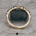 The Sapphire "Orinoco" Band, a gold ring adorned with a row of blue gemstones, is showcased standing vertically on a textured surface. The background highlights a geometric stone pattern, and the ring is partially leaning against a mossy green object. The inscription "Earth's Treasury" can be seen at the bottom.