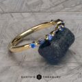 A gold bracelet adorned with blue sapphires, known as the Sapphire "Orinoco" Band, is elegantly draped over a textured dark blue stone. The background features a gray, patterned surface. The image includes the text "EARTH'S TREASURY" at the bottom center.