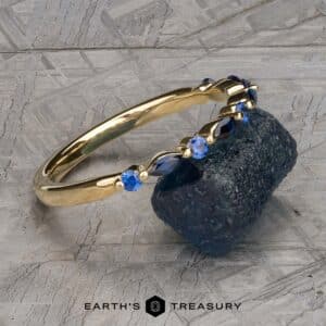 A gold bracelet adorned with blue sapphires, known as the Sapphire "Orinoco" Band, is elegantly draped over a textured dark blue stone. The background features a gray, patterned surface. The image includes the text "EARTH'S TREASURY" at the bottom center.