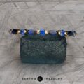 The Sapphire "Orinoco" Band is elegantly displayed on a textured blue-green stone, adorned with brilliant blue gemstones. The background features an intricate grey geometric pattern. The text at the bottom reads "Earth's Treasury.