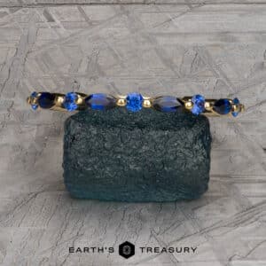 The Sapphire "Orinoco" Band is elegantly displayed on a textured blue-green stone, adorned with brilliant blue gemstones. The background features an intricate grey geometric pattern. The text at the bottom reads "Earth's Treasury.