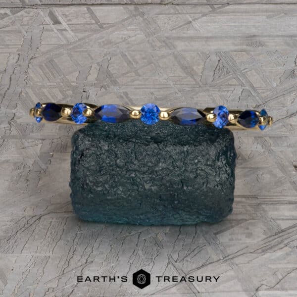 The Sapphire Orinoco Band in 18k yellow gold The Sapphire "Orinoco" Band is elegantly displayed on a textured blue-green stone, adorned with brilliant blue gemstones. The background features an intricate grey geometric pattern. The text at the bottom reads "Earth's Treasury.