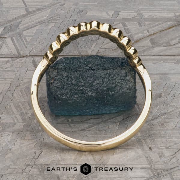 The "Taurion" Band in 18k yellow gold The "Taurion" Band, a gold ring featuring a unique scalloped edge design, is displayed upright on a flat surface with an uneven, dark blue textured rock in the center. The background has a geometric pattern, and the text "EARTH'S TREASURY" is visible at the bottom of the image.