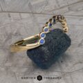 The "Taurion" Band, a gold bracelet adorned with round blue gemstones, is elegantly positioned on a textured, rough-edged stone. The bracelet features a series of evenly spaced blue gems in a curved arrangement. The background displays a pattern of intersecting gray lines, with the text "EARTH'S TREASURY" appearing at the bottom center.