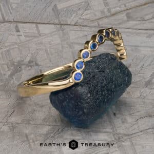 The "Taurion" Band, a gold bracelet adorned with round blue gemstones, is elegantly positioned on a textured, rough-edged stone. The bracelet features a series of evenly spaced blue gems in a curved arrangement. The background displays a pattern of intersecting gray lines, with the text "EARTH'S TREASURY" appearing at the bottom center.