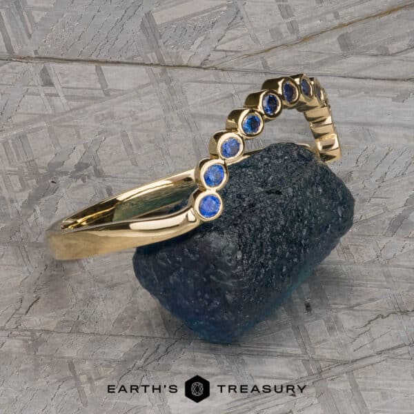 The "Taurion" Band in 18k yellow gold The "Taurion" Band, a gold bracelet adorned with round blue gemstones, is elegantly positioned on a textured, rough-edged stone. The bracelet features a series of evenly spaced blue gems in a curved arrangement. The background displays a pattern of intersecting gray lines, with the text "EARTH'S TREASURY" appearing at the bottom center.