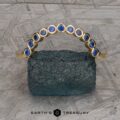 The "Taurion" Band, a gold ring adorned with a graceful curve of blue gemstones, is elegantly showcased on a textured, rough stone. The background features an intricately patterned gray metallic surface with the words "EARTH'S TREASURY" visible at the bottom of the image.