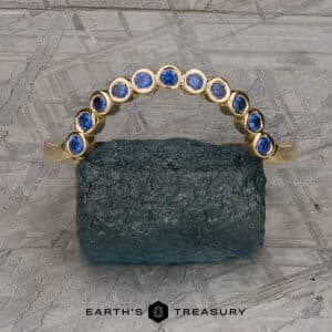 The "Taurion" Band, a gold ring adorned with a graceful curve of blue gemstones, is elegantly showcased on a textured, rough stone. The background features an intricately patterned gray metallic surface with the words "EARTH'S TREASURY" visible at the bottom of the image.
