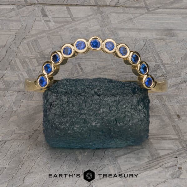 The "Taurion" Band in 18k yellow gold The "Taurion" Band, a gold ring adorned with a graceful curve of blue gemstones, is elegantly showcased on a textured, rough stone. The background features an intricately patterned gray metallic surface with the words "EARTH'S TREASURY" visible at the bottom of the image.