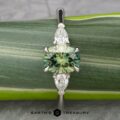 Close-up of The Cathedral “Merope” Three-Stone Ring showcasing a central emerald-cut green gemstone flanked by two smaller teardrop-shaped clear stones on either side. The ring is set against a green leaf background with the logo and text "EARTH'S TREASURY" visible at the bottom.