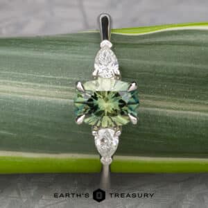 Close-up of The Cathedral “Merope” Three-Stone Ring showcasing a central emerald-cut green gemstone flanked by two smaller teardrop-shaped clear stones on either side. The ring is set against a green leaf background with the logo and text "EARTH'S TREASURY" visible at the bottom.