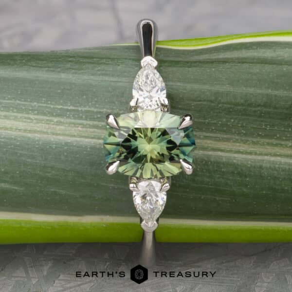 The cathedral Merope ring in platinum with 1.50-carat Australian sapphire Close-up of The Cathedral “Merope” Three-Stone Ring showcasing a central emerald-cut green gemstone flanked by two smaller teardrop-shaped clear stones on either side. The ring is set against a green leaf background with the logo and text "EARTH'S TREASURY" visible at the bottom.
