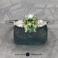 The Cathedral “Merope” Three-Stone Ring, showcasing a green gemstone flanked by two smaller white stones, is displayed on a textured dark green surface. The text "Earth's Treasury" is printed below the ring.