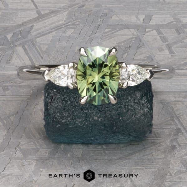 The cathedral Merope ring in platinum with 1.50-carat Australian sapphire The Cathedral “Merope” Three-Stone Ring, showcasing a green gemstone flanked by two smaller white stones, is displayed on a textured dark green surface. The text "Earth's Treasury" is printed below the ring.
