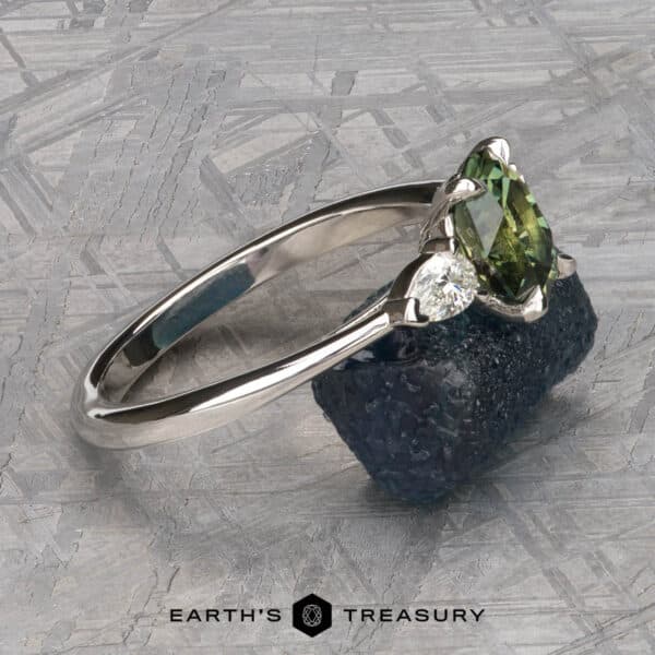 The cathedral Merope ring in platinum with 1.50-carat Australian sapphire The Cathedral “Merope” Three-Stone Ring, with its striking silver band, showcases a central oval green gemstone flanked by a smaller round white gemstone. It is artfully displayed on rugged dark blue stone against a textured grey backdrop, with the words "Earth's Treasury" elegantly inscribed at the bottom.