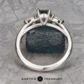 The Cathedral “Merope” Three-Stone Ring, adorned with a dark green gemstone, is highlighted against a textured green stone background. The backdrop features geometric patterns, infusing a modern flair into the presentation. The image is branded with the "Earth's Treasury" logo at the bottom.