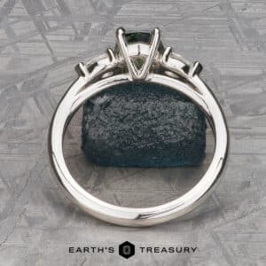 The Cathedral “Merope” Three-Stone Ring, adorned with a dark green gemstone, is highlighted against a textured green stone background. The backdrop features geometric patterns, infusing a modern flair into the presentation. The image is branded with the "Earth's Treasury" logo at the bottom.