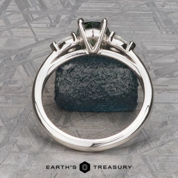 The cathedral Merope ring in platinum with 1.50-carat Australian sapphire The Cathedral “Merope” Three-Stone Ring, adorned with a dark green gemstone, is highlighted against a textured green stone background. The backdrop features geometric patterns, infusing a modern flair into the presentation. The image is branded with the "Earth's Treasury" logo at the bottom.