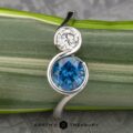Introducing The “Nerissa” Two-Stone Ring, showcasing a harmonious blend of a round white diamond and a larger blue gemstone, elegantly set in a sleek silver swirl design. Each ring is carefully photographed against a green and white striped leaf with the Earth's Treasury logo proudly displayed below.