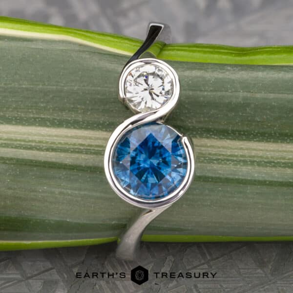 The Nerissa ring in platinum with 1.77-carat Montana sapphire and 4.6mm heirloom diamond Introducing The “Nerissa” Two-Stone Ring, showcasing a harmonious blend of a round white diamond and a larger blue gemstone, elegantly set in a sleek silver swirl design. Each ring is carefully photographed against a green and white striped leaf with the Earth's Treasury logo proudly displayed below.