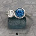 The "Nerissa" Two-Stone Ring, crafted in silver with a large round blue gemstone and a smaller clear gemstone beside it, is elegantly displayed on a textured teal rock. A veined stone pattern graces the background, with the text "EARTH'S TREASURY" appearing at the bottom.