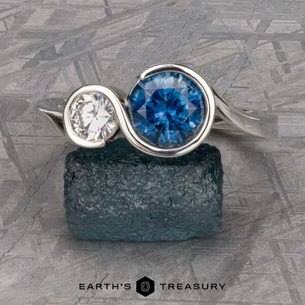 The Nerissa ring in platinum with 1.77-carat Montana sapphire and 4.6mm heirloom diamond The "Nerissa" Two-Stone Ring, crafted in silver with a large round blue gemstone and a smaller clear gemstone beside it, is elegantly displayed on a textured teal rock. A veined stone pattern graces the background, with the text "EARTH'S TREASURY" appearing at the bottom.