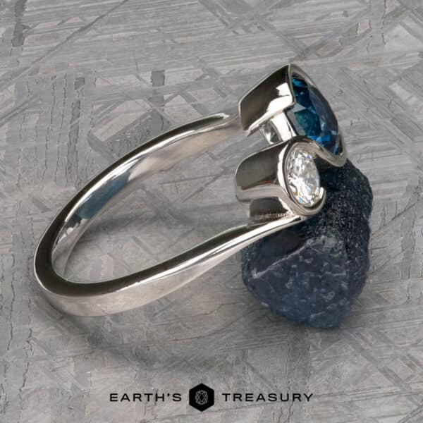 The Nerissa ring in platinum with 1.77-carat Montana sapphire and 4.6mm heirloom diamond The “Nerissa” Two-Stone Ring, a silver open-band piece featuring a polished blue gemstone and a clear diamond, rests against a rough blue mineral on a textured gray background. The text "Earth's Treasury" is at the bottom of the image.