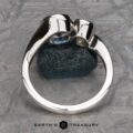 The "Nerissa" Two-Stone Ring, crafted in silver and adorned with a blue gemstone, is prominently displayed upright on a green textured stone. The background showcases a geometric pattern, with the text "EARTH'S TREASURY" visible at the bottom center of the image.