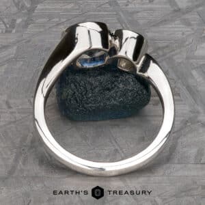 The "Nerissa" Two-Stone Ring, crafted in silver and adorned with a blue gemstone, is prominently displayed upright on a green textured stone. The background showcases a geometric pattern, with the text "EARTH'S TREASURY" visible at the bottom center of the image.