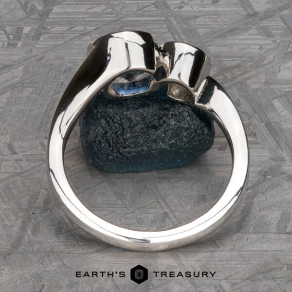 The Nerissa ring in platinum with 1.77-carat Montana sapphire and 4.6mm heirloom diamond The "Nerissa" Two-Stone Ring, crafted in silver and adorned with a blue gemstone, is prominently displayed upright on a green textured stone. The background showcases a geometric pattern, with the text "EARTH'S TREASURY" visible at the bottom center of the image.