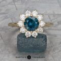 The "Theodora" ring in 14k yellow gold showcases a striking 1.84-carat teal Montana gemstone at its center, surrounded by sparkling diamonds, all beautifully set against a textured grey backdrop.
