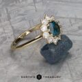 The breathtaking "Theodora" ring, set in 14k yellow gold, showcases a 1.84-carat Montana sapphire at its heart, surrounded by dazzling diamonds, elegantly presented on a textured backdrop with a rough blue stone.