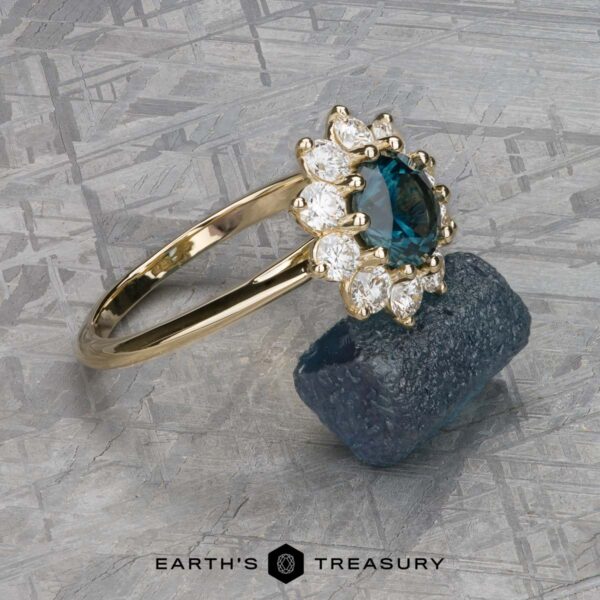 The "Theodora" ring in 14k yellow gold with 1.84-carat Montana sapphire The breathtaking "Theodora" ring, set in 14k yellow gold, showcases a 1.84-carat Montana sapphire at its heart, surrounded by dazzling diamonds, elegantly presented on a textured backdrop with a rough blue stone.