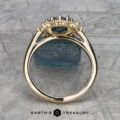 The "Theodora" ring in 14k yellow gold boasts a 1.84-carat teal Montana sapphire, elegantly accented with diamonds and displayed against a textured blue mineral background.