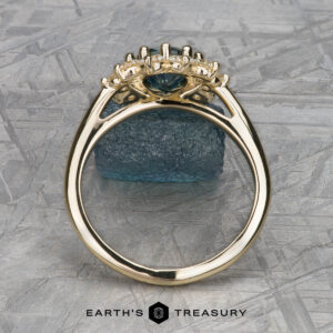 The "Theodora" ring in 14k yellow gold boasts a 1.84-carat teal Montana sapphire, elegantly accented with diamonds and displayed against a textured blue mineral background.