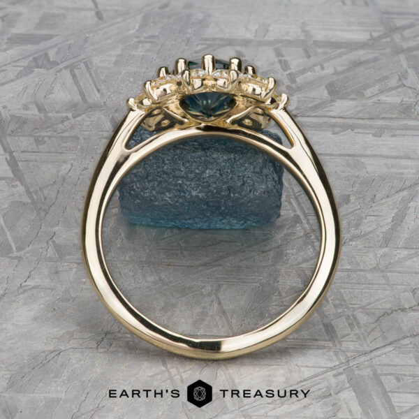 The "Theodora" ring in 14k yellow gold with 1.84-carat Montana sapphire The "Theodora" ring in 14k yellow gold boasts a 1.84-carat teal Montana sapphire, elegantly accented with diamonds and displayed against a textured blue mineral background.
