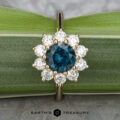 The "Theodora" ring in 14k yellow gold showcases a captivating 1.84-carat blue central gemstone, elegantly encircled by small white gems, all set against a backdrop of lush green leaves. The text at the bottom, "Earth's Treasury," encapsulates the essence of exquisite jewelry reminiscent of Theodora's timeless allure.
