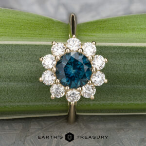 The "Theodora" ring in 14k yellow gold showcases a captivating 1.84-carat blue central gemstone, elegantly encircled by small white gems, all set against a backdrop of lush green leaves. The text at the bottom, "Earth's Treasury," encapsulates the essence of exquisite jewelry reminiscent of Theodora's timeless allure.