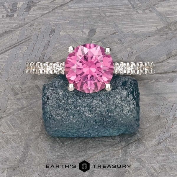The classic Blumia ring in 18k white gold with 1.72-carat Montana sapphire Introducing The Classic Pave "Blumia" Ring, a stunning piece featuring a vibrant pink gemstone set in a silver band adorned with small sparkling diamonds. Displayed on a textured blue stone, this exquisite ring is showcased with the logo "Earth's Treasury" beneath it.