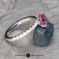 The Classic Pave "Blumia" Ring, featuring a marquise-shaped pink gemstone set in silver and adorned with small clear stones around the band, is displayed on a blue rock against a textured metallic background. The text "Earth's Treasury" is visible at the bottom of the image.